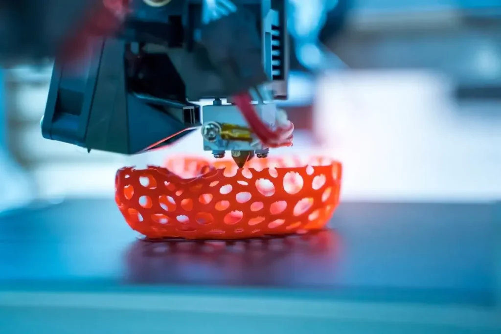 Блог пост - Еволюцията на 3D технологията и изкуството.Крупен план на 3D принтер, който печата обект с мрежеста структура в яркочервен цвят. Дюзата на принтера депозира материала слой по слой върху печатащата платформа. Фонът е размазан, с преобладаващи сини и бели тонове, създавайки технологична и футуристична атмосфера.